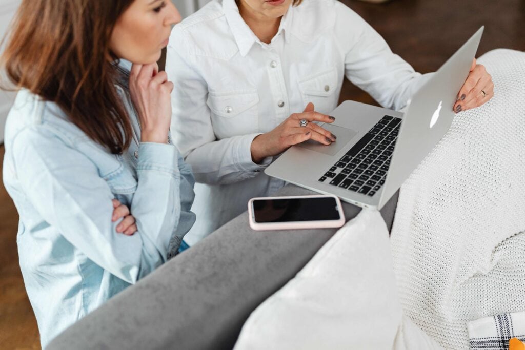 Two women working together on a laptop in a cozy indoor setting with a smartphone nearby